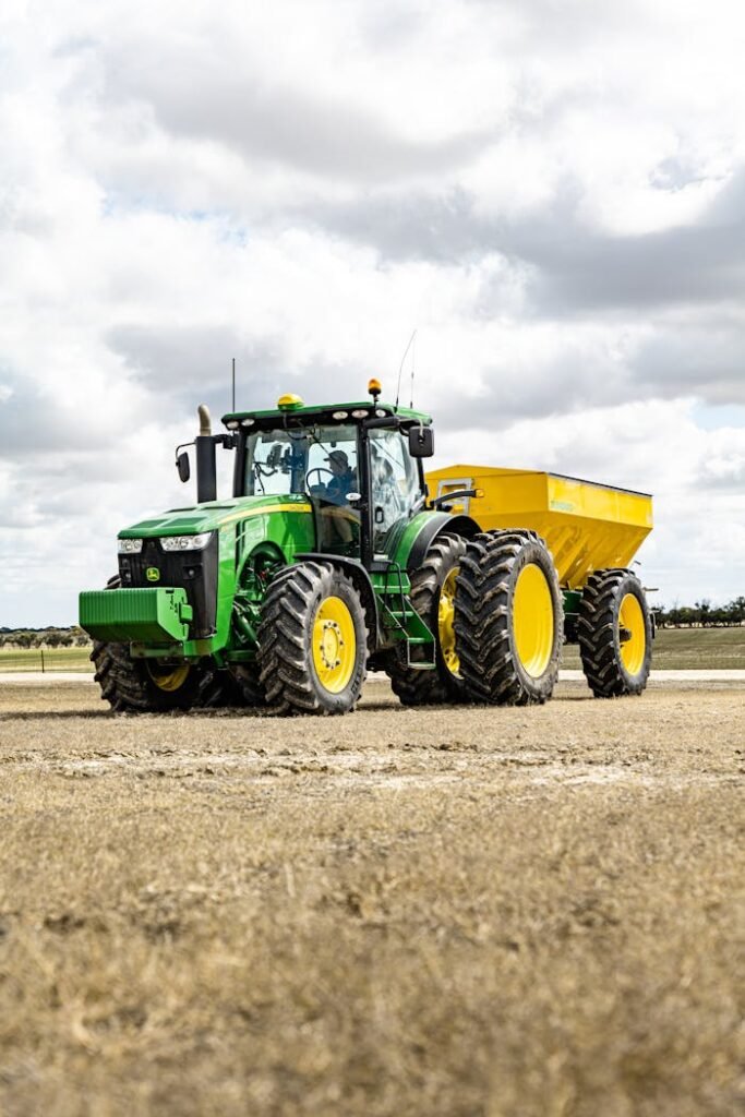 Contemporary agricultural machinery with trailer riding along grassy field in countryside against cloudy sky